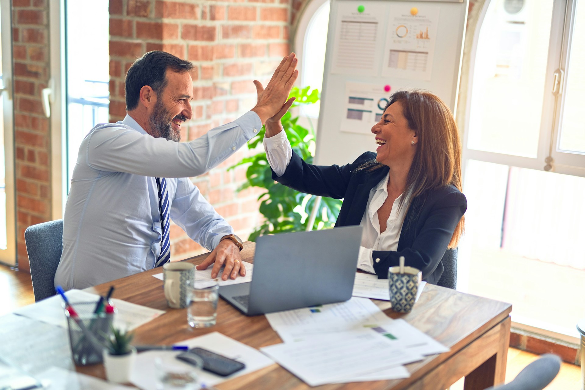 Businessman and business woman giving high fives at a table with a laptop in a brightly lit room